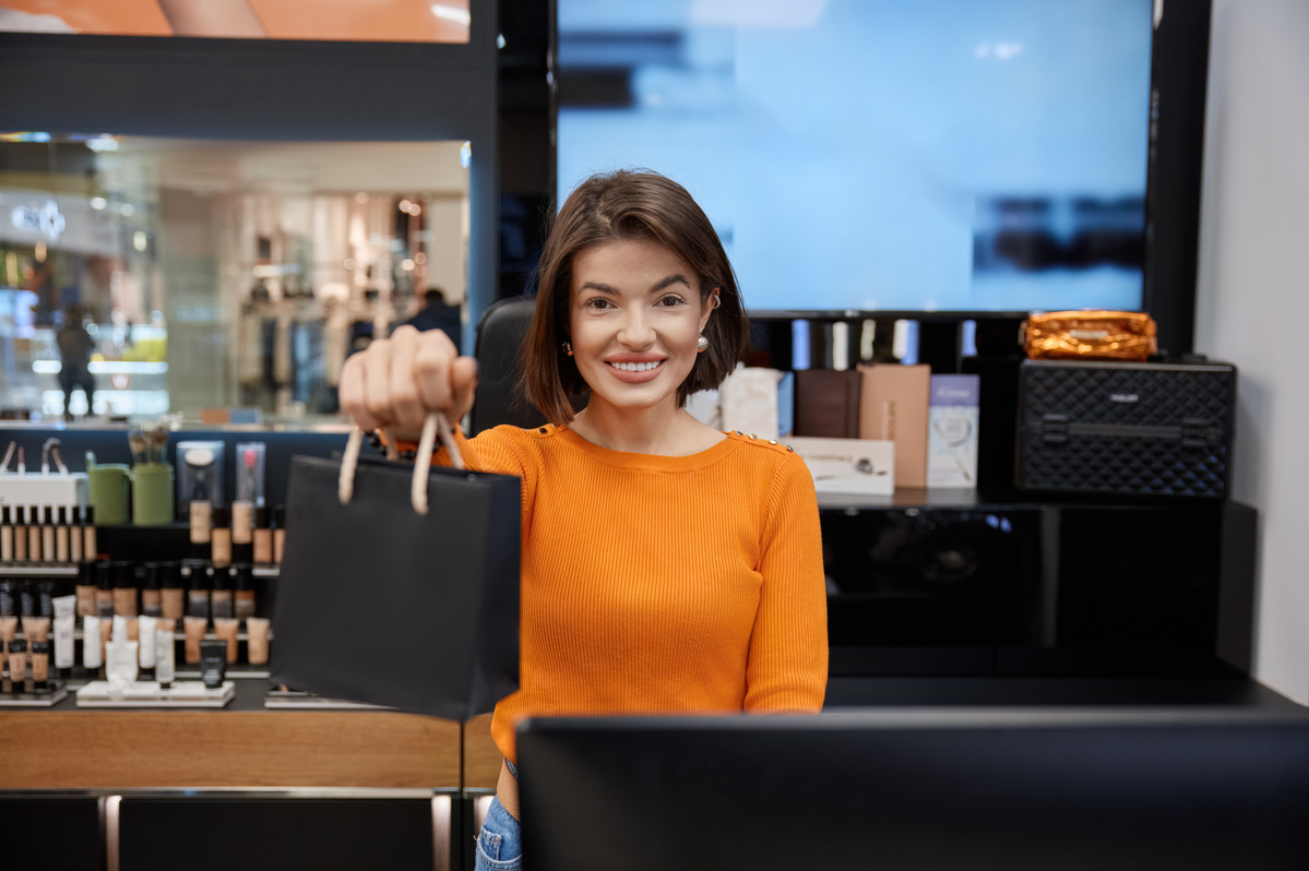 Beautiful smiling saleswoman in cosmetics store holding out the package. Attractive positive young lady working with customers